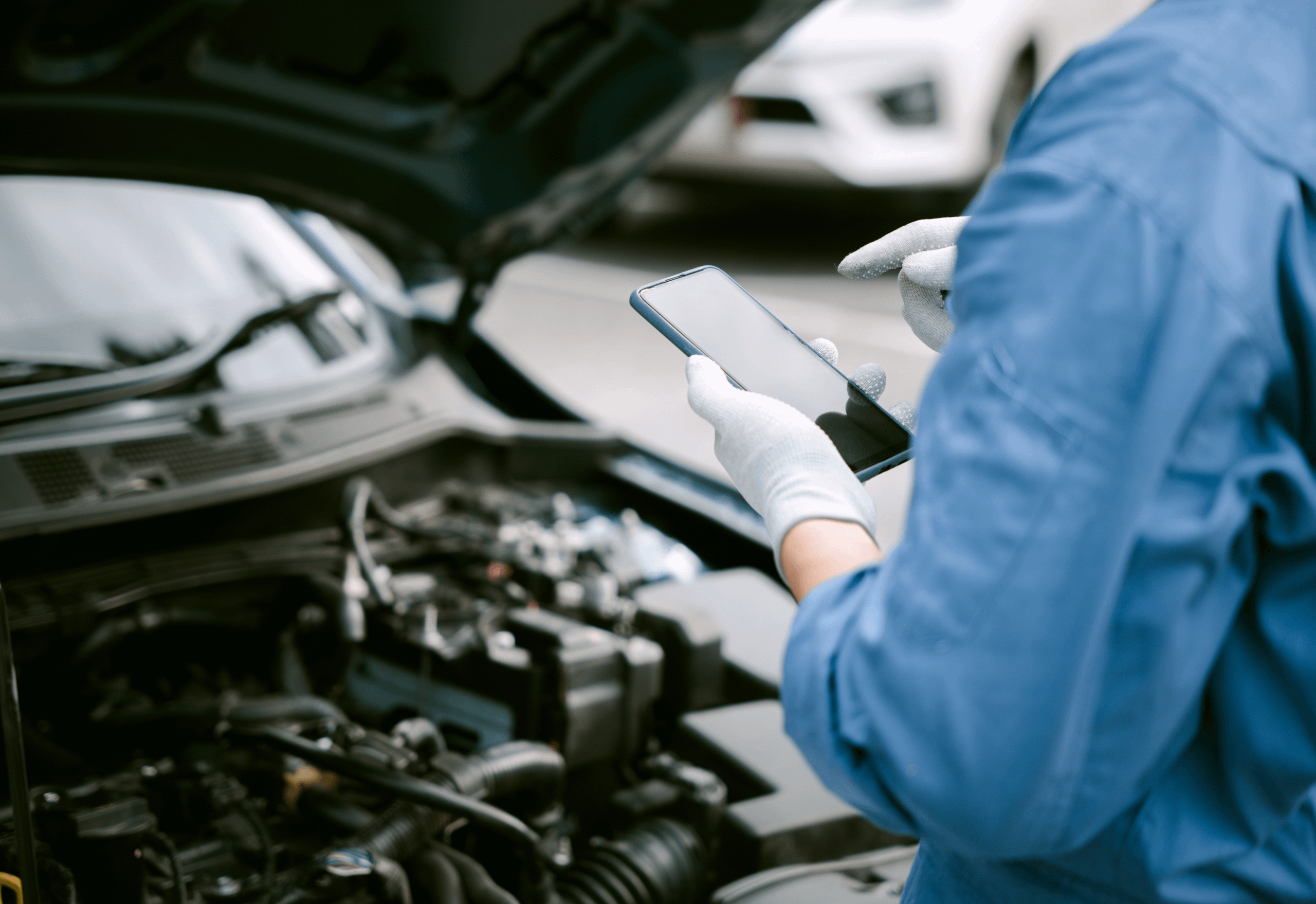 Mechanic in blue overalls and white gloves stood in front of car with the bonnet up, holding a phone.
