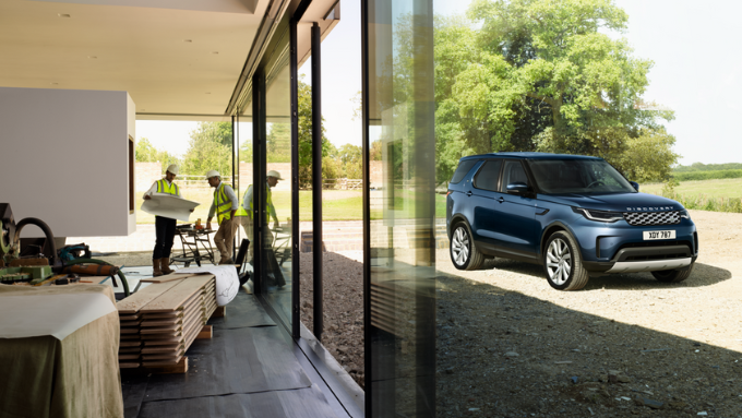 A blue Land Rover Discovery parked outside a modern house while construction workers work inside near large glass doors.