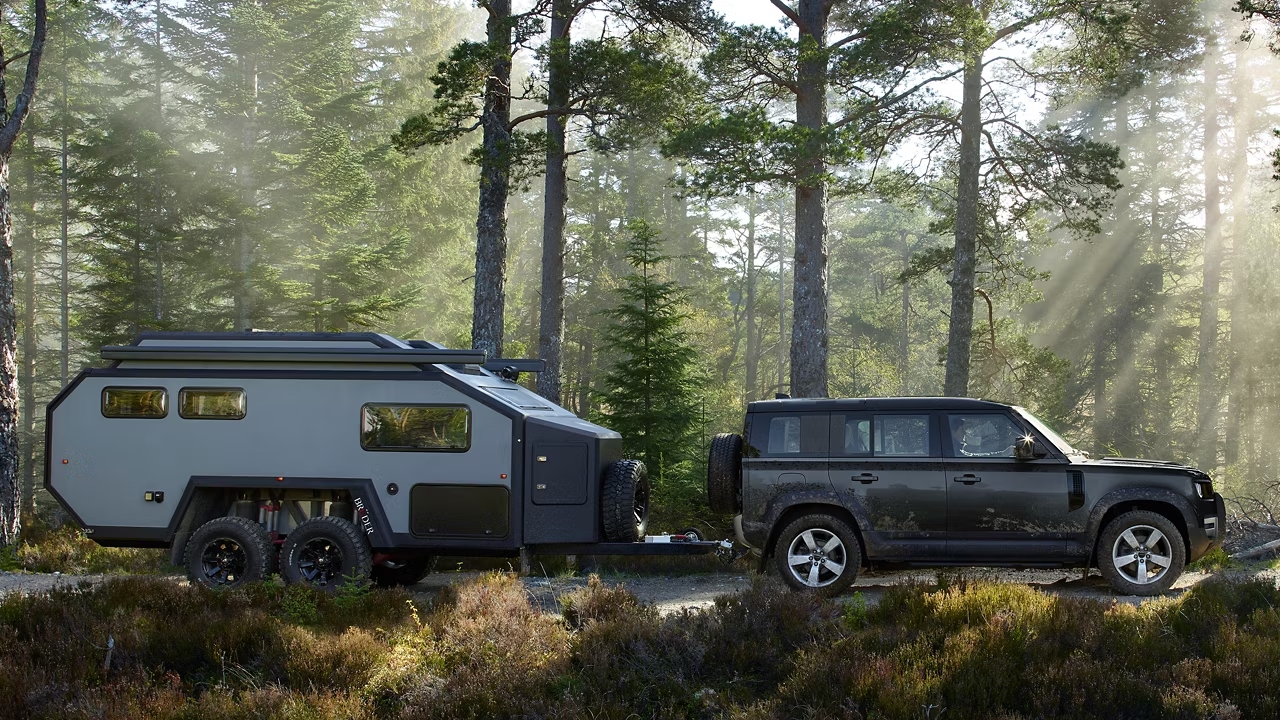 Defender 110 towing an off-road camper trailer through a forest clearing.