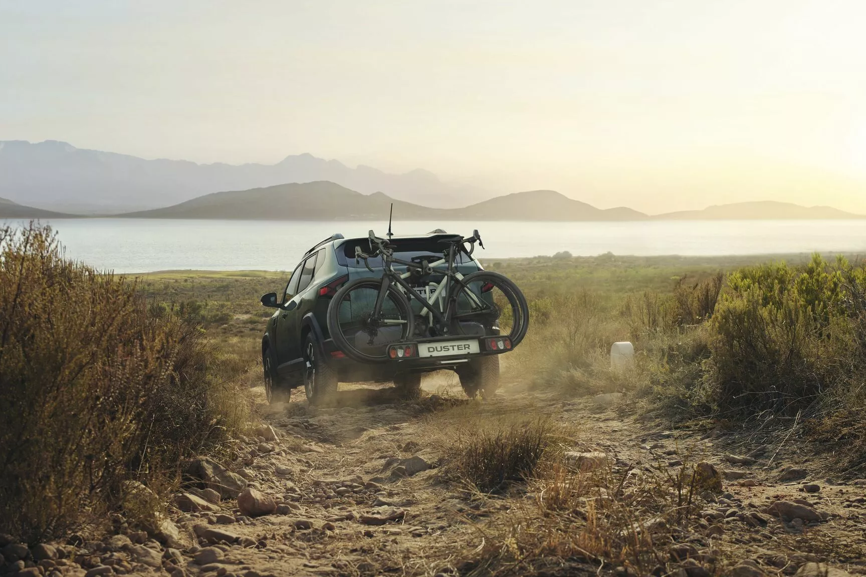 Dacia Duster driving along a rocky dirt track with two bicycles mounted on the rear, overlooking a lake and distant mountains at sunset.