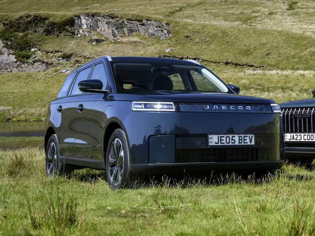 Front three-quarter view of the Jaecoo E5 electric SUV in deep blue, featuring a distinctive full-width LED light bar, bold grille design and multi-spoke alloy wheels set against rugged countryside scenery.