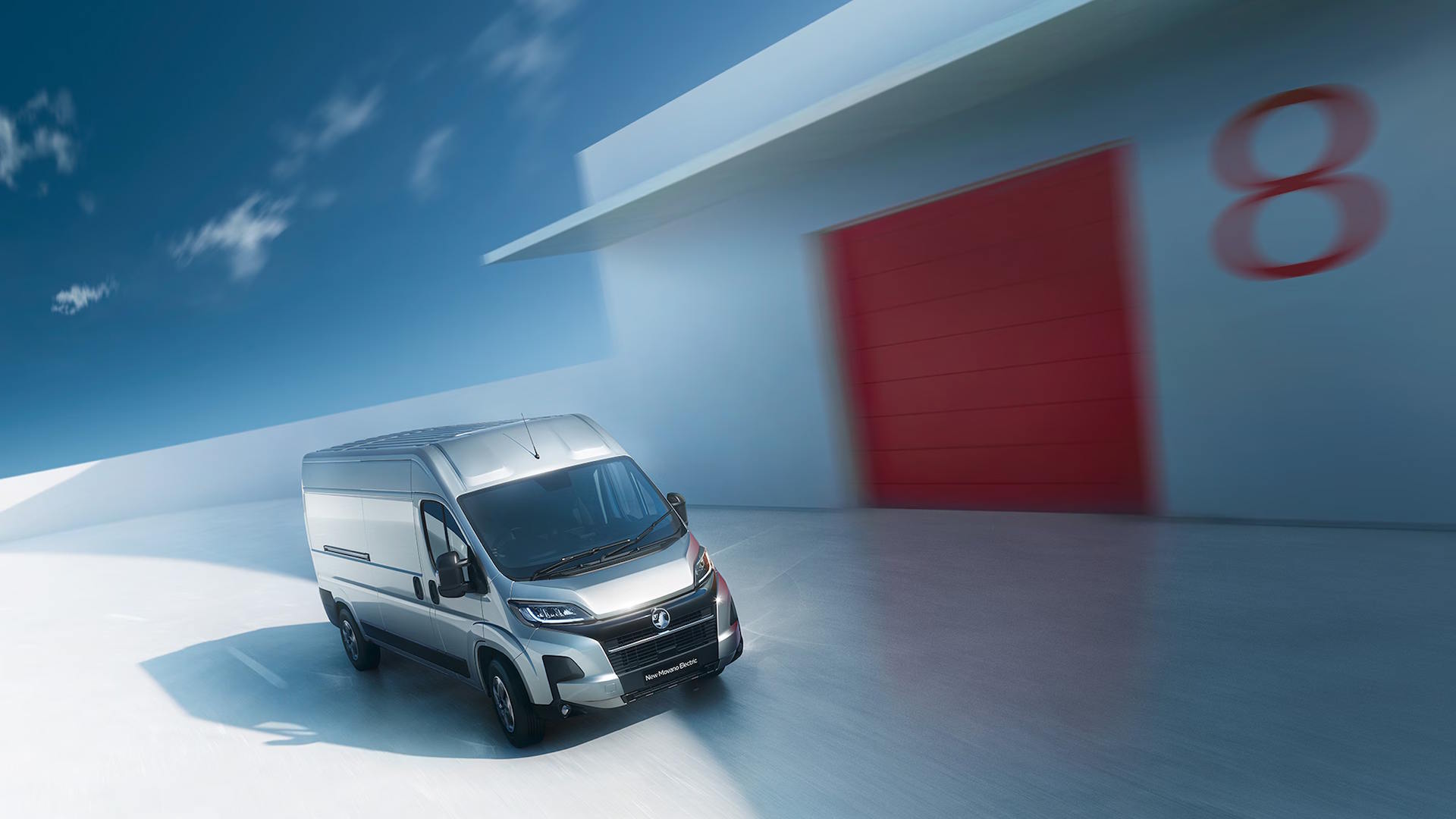 Grey Vauxhall Movano driving through a bright studio setting beside a red door.