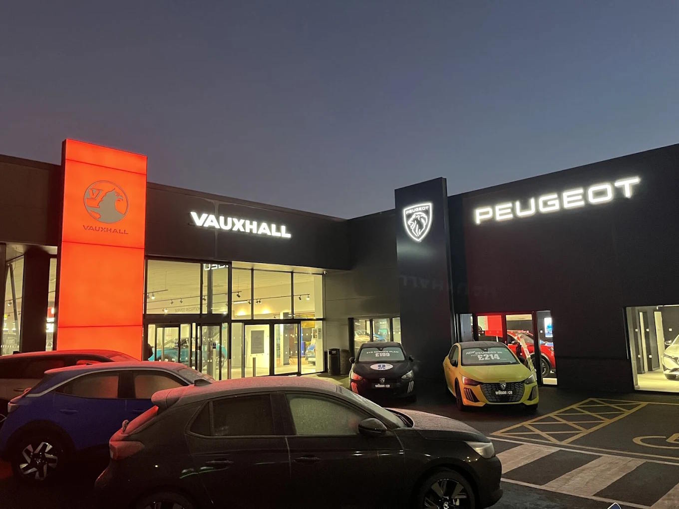 Exterior twilight view of Peugeot Bolton dealership, featuring illuminated Peugeot and Vauxhall signage, modern showroom frontage and forecourt displaying new Peugeot models.