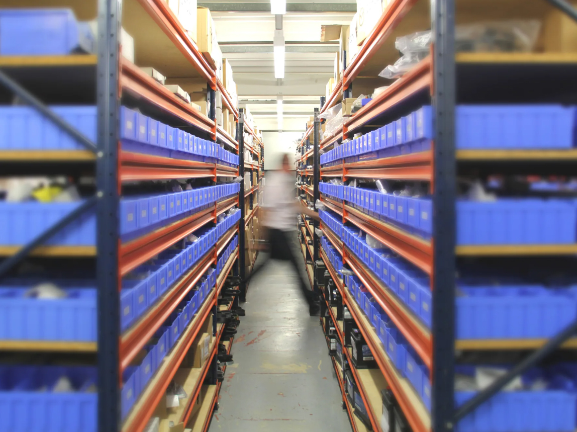 Central aisle view of a dealership parts department featuring rows of blue component bins on orange racking, with staff member accessing stock.