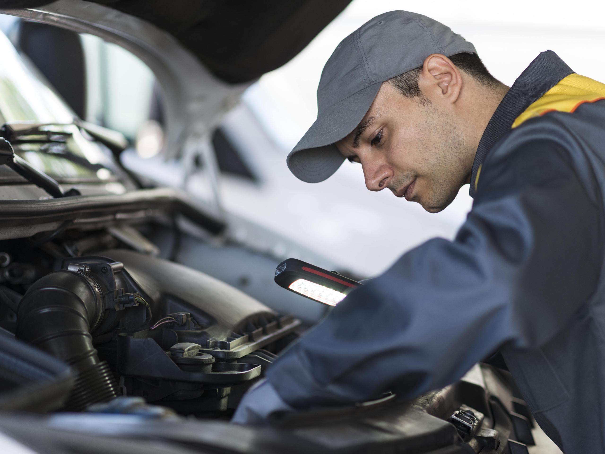 Vehicle technician inspecting an engine bay with a handheld work light in an automotive workshop.
