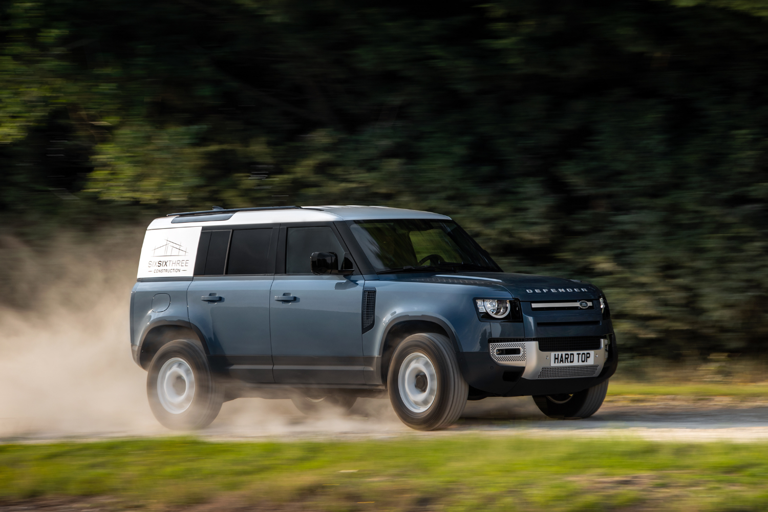 A Land Rover Defender Hard Top driving on a dirt road, kicking up dust with trees in the background.