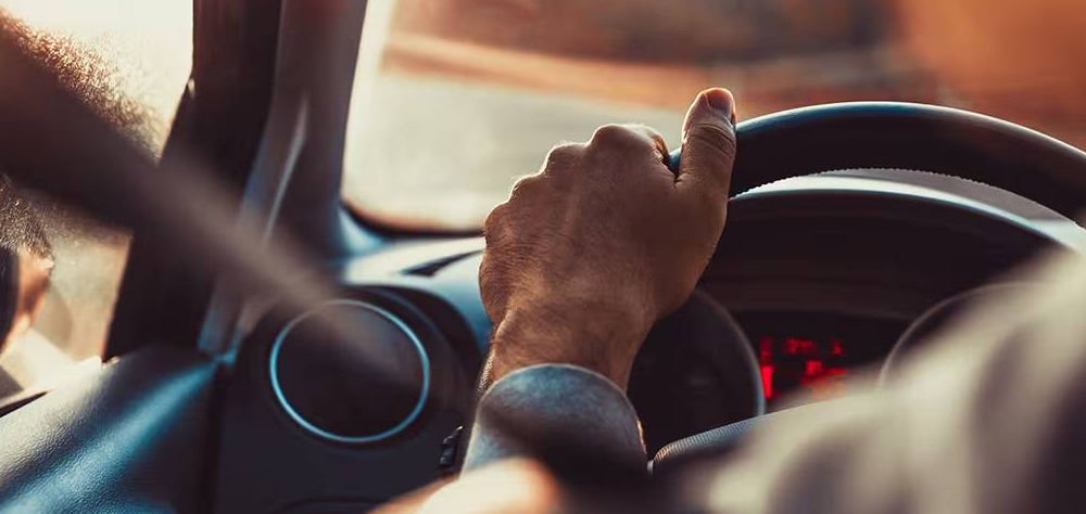 Close-up of a driver's hand gripping the steering wheel while driving, with sunlight streaming through the windscreen