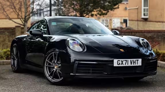 Black Porsche 911 coupe with dark alloy wheels, parked on tarmac in front of houses and trees, viewed from the front three‑quarter angle.