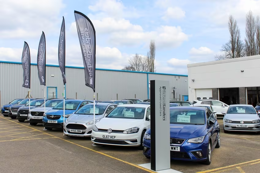 Forecourt display of approved used cars at dealership with multiple vehicles parked in rows and promotional flags visible.