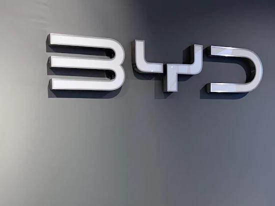 BYD logo displayed on a grey dealership wall, featuring polished chrome lettering with the distinctive three-character brand emblem