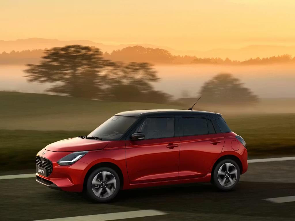 Side profile of the Suzuki Swift supermini in vibrant red with contrasting black roof, showcasing its compact proportions, angular LED headlights and sporty alloy wheels on a misty country road.