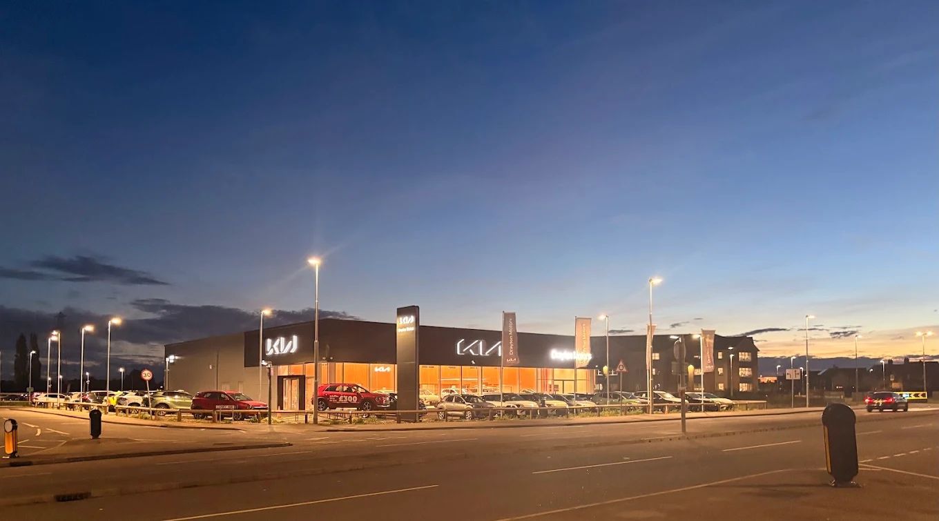 Boston Kia dealership exterior at dusk, featuring illuminated showroom with floor-to-ceiling windows, prominent Kia signage and forecourt display vehicles under dramatic twilight sky.