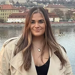 Person standing beside a river with historic buildings visible in the background.