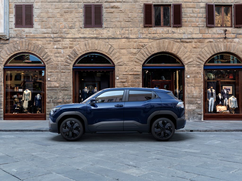 Side profile view of the Suzuki e Vitara compact SUV in dark blue, featuring rugged black plastic cladding, gloss black alloy wheels and a bold crossover stance.
