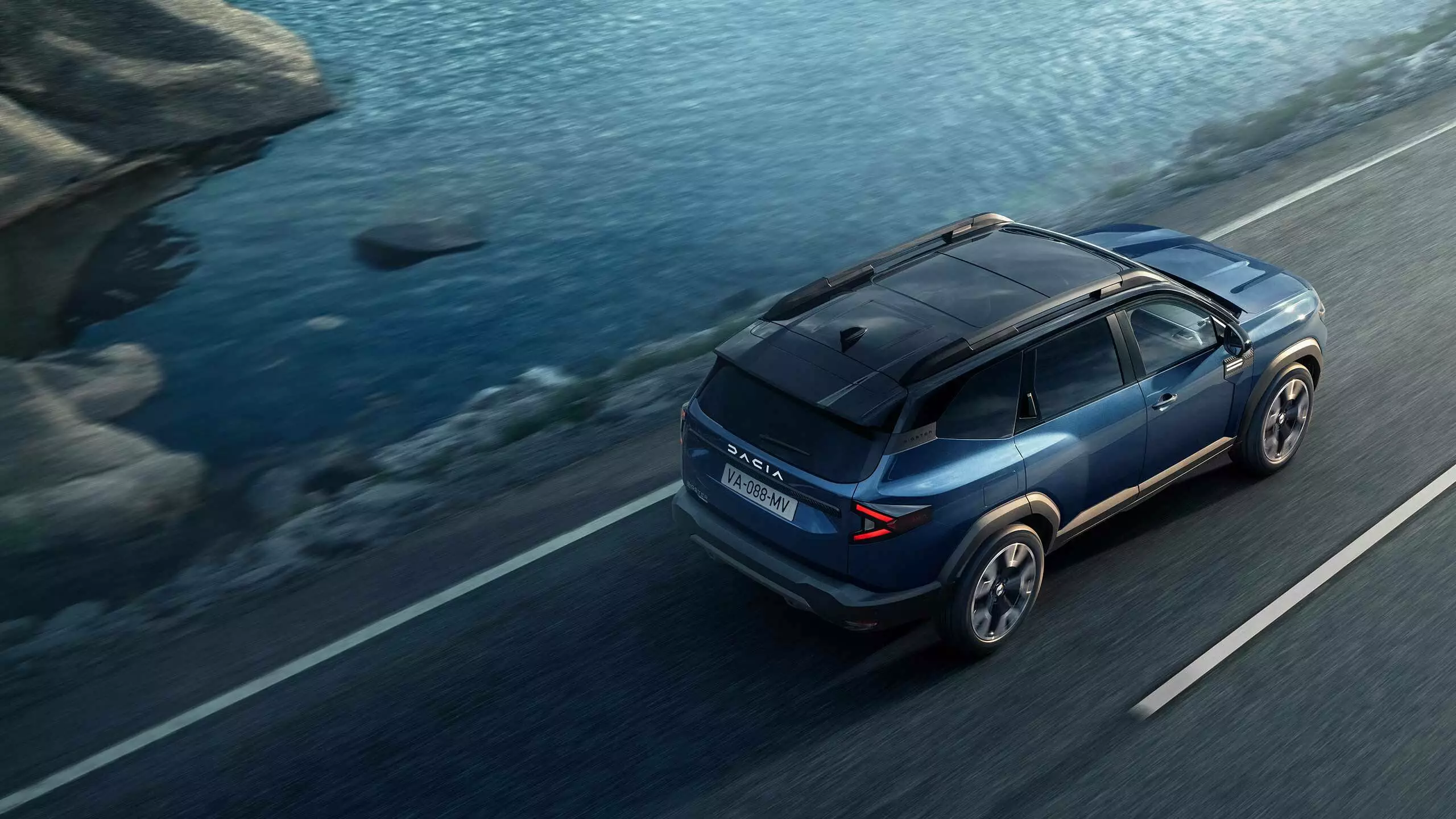 Dacia Jogger driving along a coastal road beside calm blue water, viewed from above with rocky shoreline alongside.