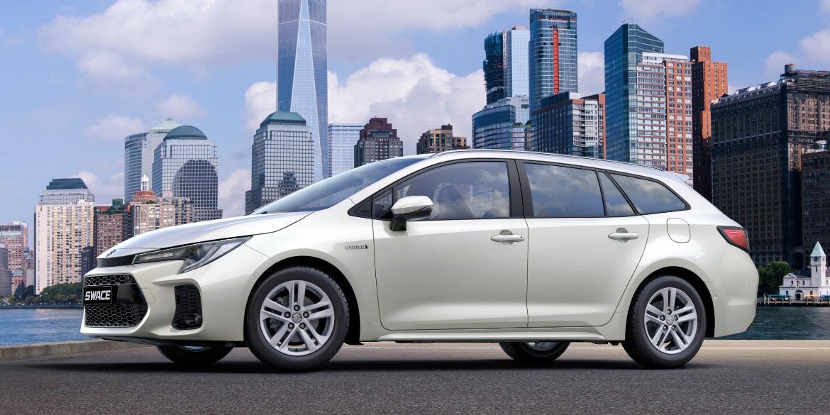 Side profile view of the Suzuki Swace estate in pearl white, showcasing its sleek silhouette, multi-spoke alloy wheels and aerodynamic roofline against a city skyline backdrop.