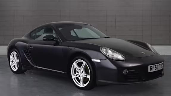 Dark grey Porsche Cayman coupe parked in a studio with a tiled grey wall background, shown from the front three‑quarter view.