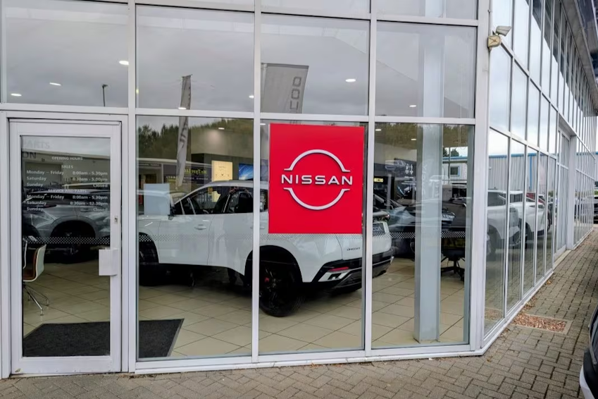 Exterior view through glass of Nissan showroom entrance with Nissan logo signage and vehicle displayed inside.