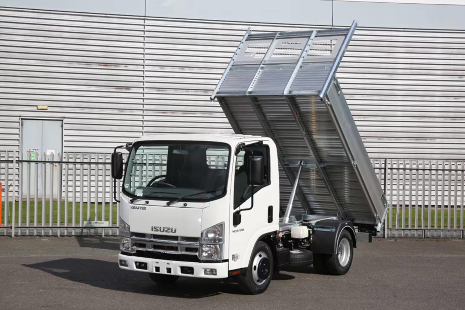 White Isuzu N55 150 chassis cab tipper truck with dump bed partially lifted, positioned outside a warehouse.