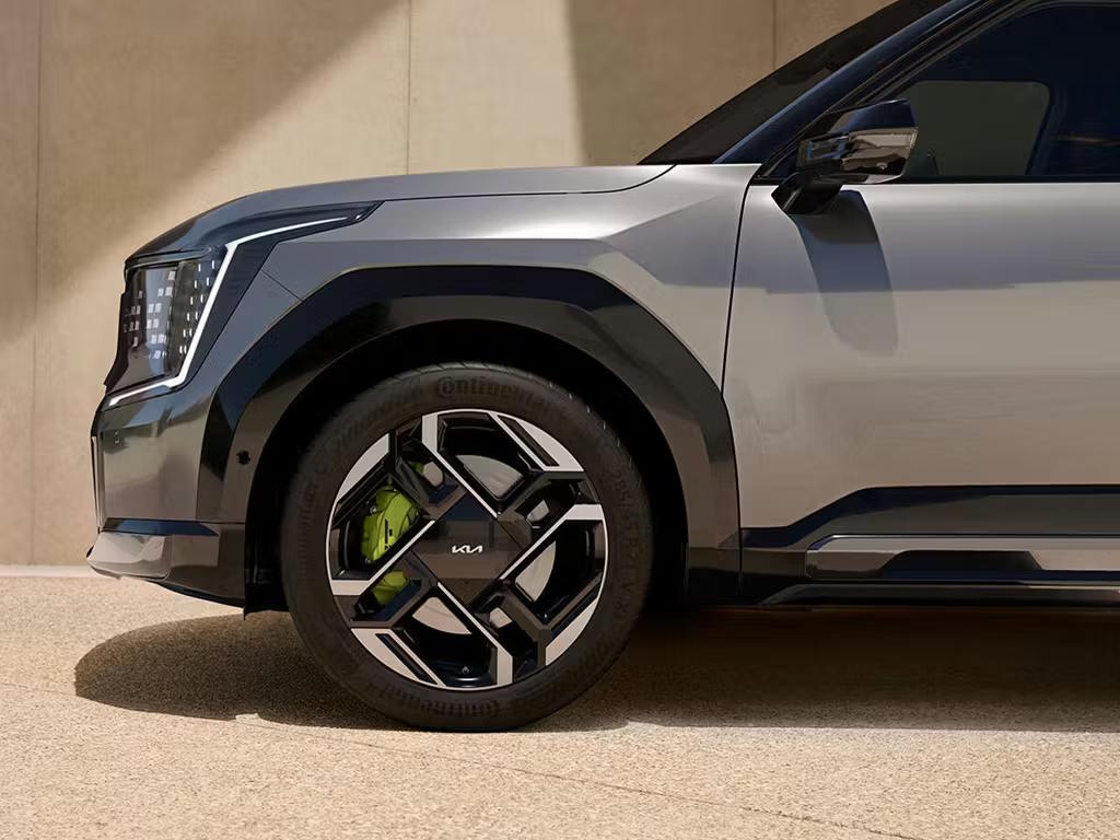 Front three-quarter close-up of the Kia EV9 GT in grey, showcasing its distinctive geometric LED headlights, bold alloy wheel design with lime green brake callipers and muscular wheel arches.