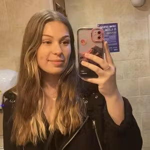 Person taking a mirror selfie while wearing a black jacket in a tiled interior.