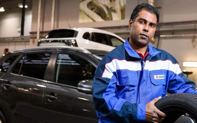 Suzuki technician in blue workshop uniform holding a tyre in front of a dark Suzuki car inside a service bay.