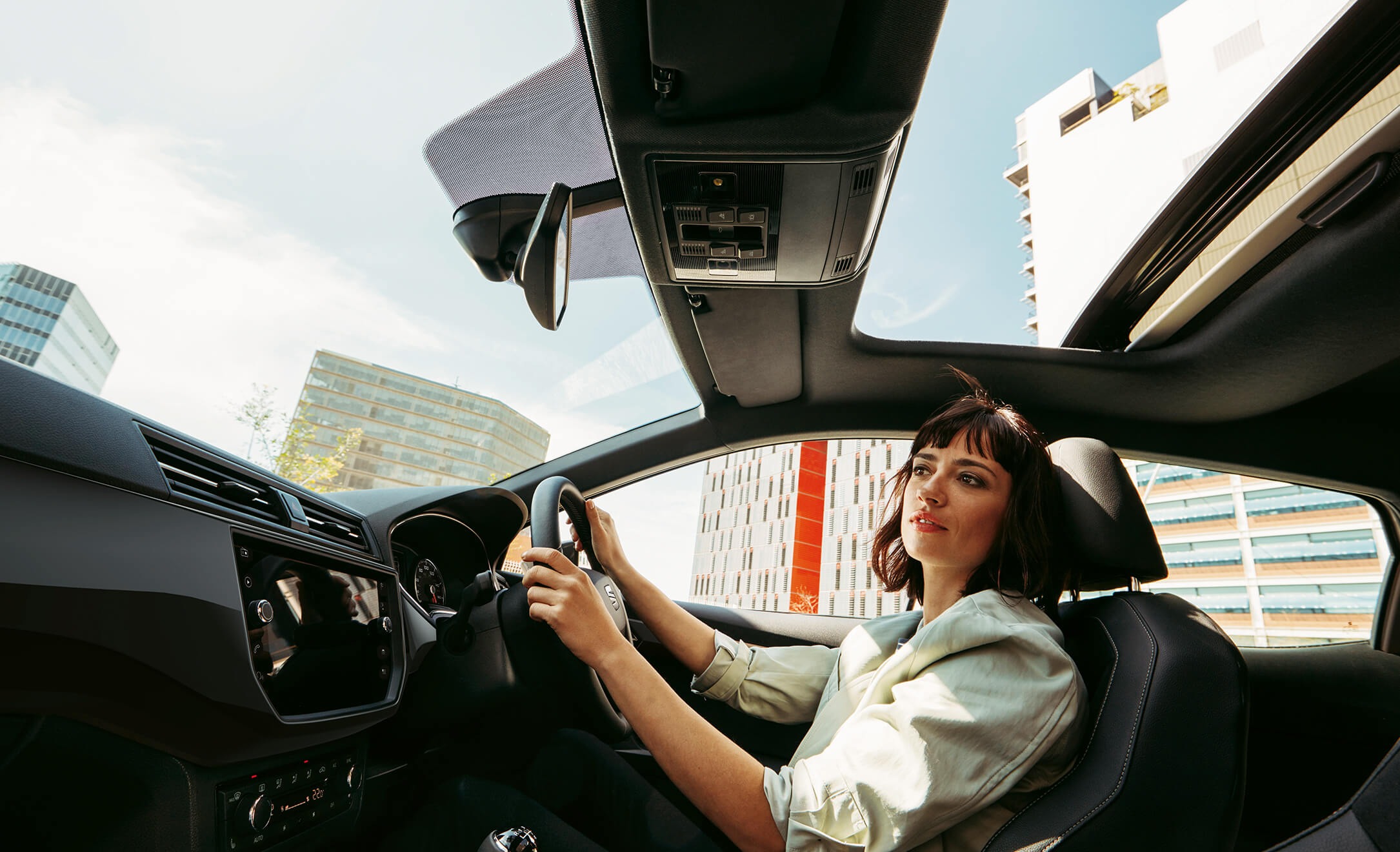 Woman driving a SEAT with the sunroof open in an urban setting.