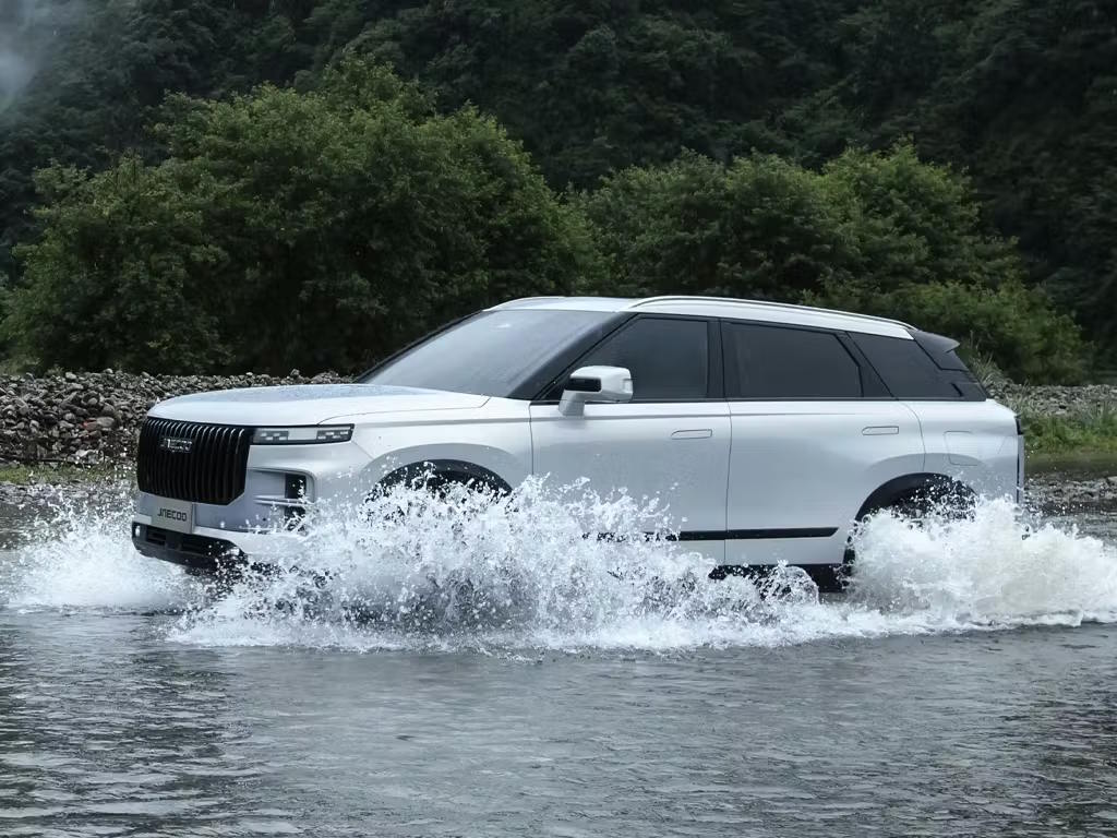 White Jaecoo 7 SUV powering through a river crossing, showcasing its off-road capability with water splashing dramatically around the bold front grille and wheel arches.