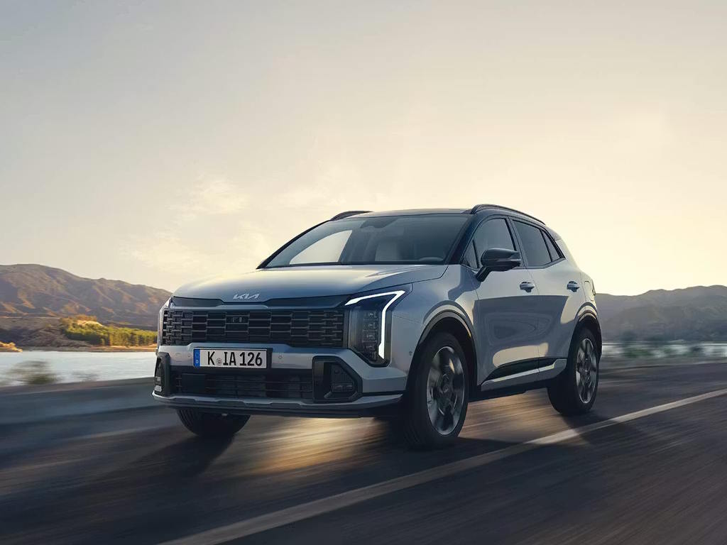 Front three-quarter view of the Kia Sportage SUV in motion, featuring the bold tiger-nose grille, distinctive boomerang LED headlights and sculpted grey metallic bodywork against a mountainous backdrop.
