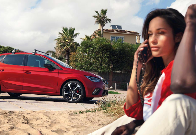 Red Seat Ibiza parked near a sandy beach with a woman sitting in the foreground.