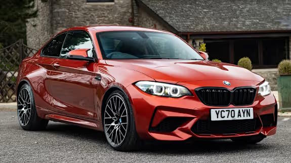 Metallic red BMW M2 coupe with aggressive front bumper and multi‑spoke alloy wheels, parked on a driveway in front of stone buildings.