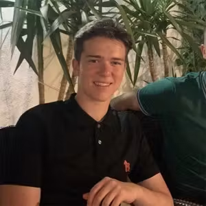 Person seated indoors smiling towards the camera with plants in the background.