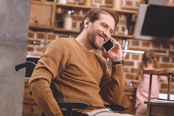 Man in a wheelchair smiling while talking on the phone in a warm, home setting.
