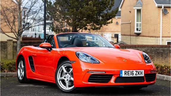 Bright orange Porsche 718 Boxster convertible parked on tarmac in front of houses and trees, viewed from the front three‑quarter angle.