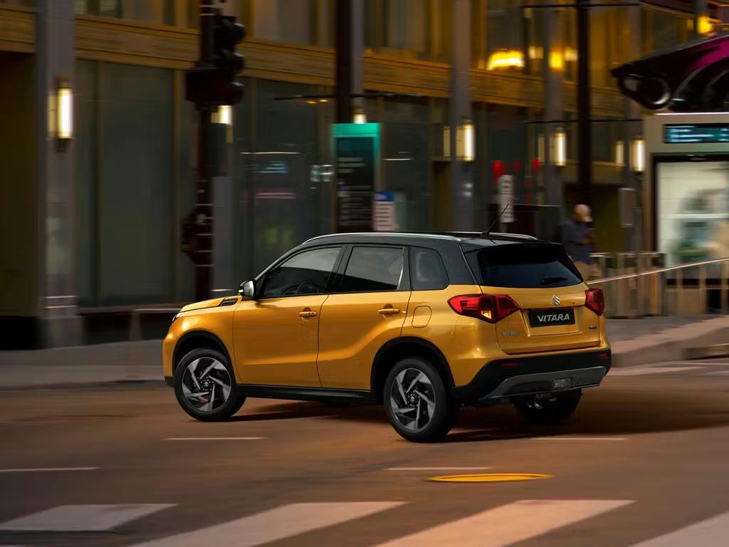 Rear three-quarter view of the Suzuki Vitara compact SUV in vibrant yellow with contrasting black roof, showcasing distinctive rear light clusters and bold wheel arch cladding in an urban setting.