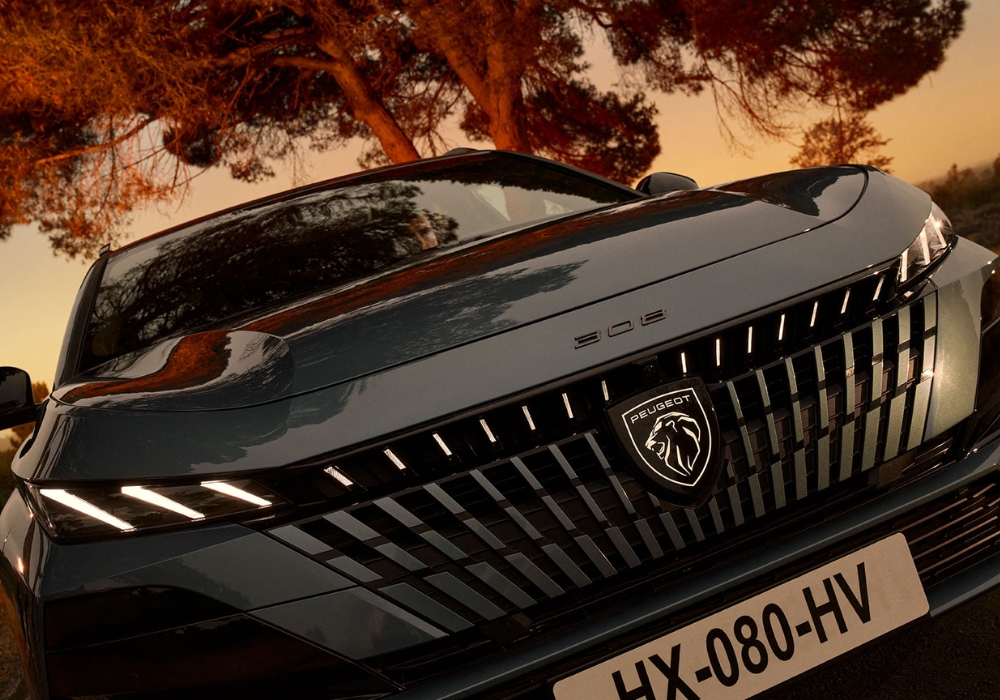 Close-up of Peugeot 308 SW front grille and badge with LED headlights illuminated at sunset.