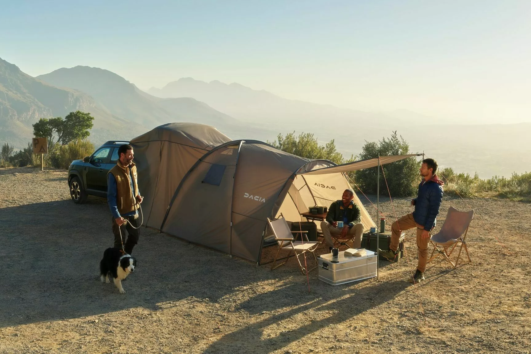 Dacia Duster parked beside a campsite with a large tent and awning, as three people relax outdoors with a dog in a mountainous landscape.