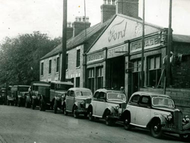 Vintage Ford garage with a line of parked cars and delivery vehicles from the mid-20th century on a street.
