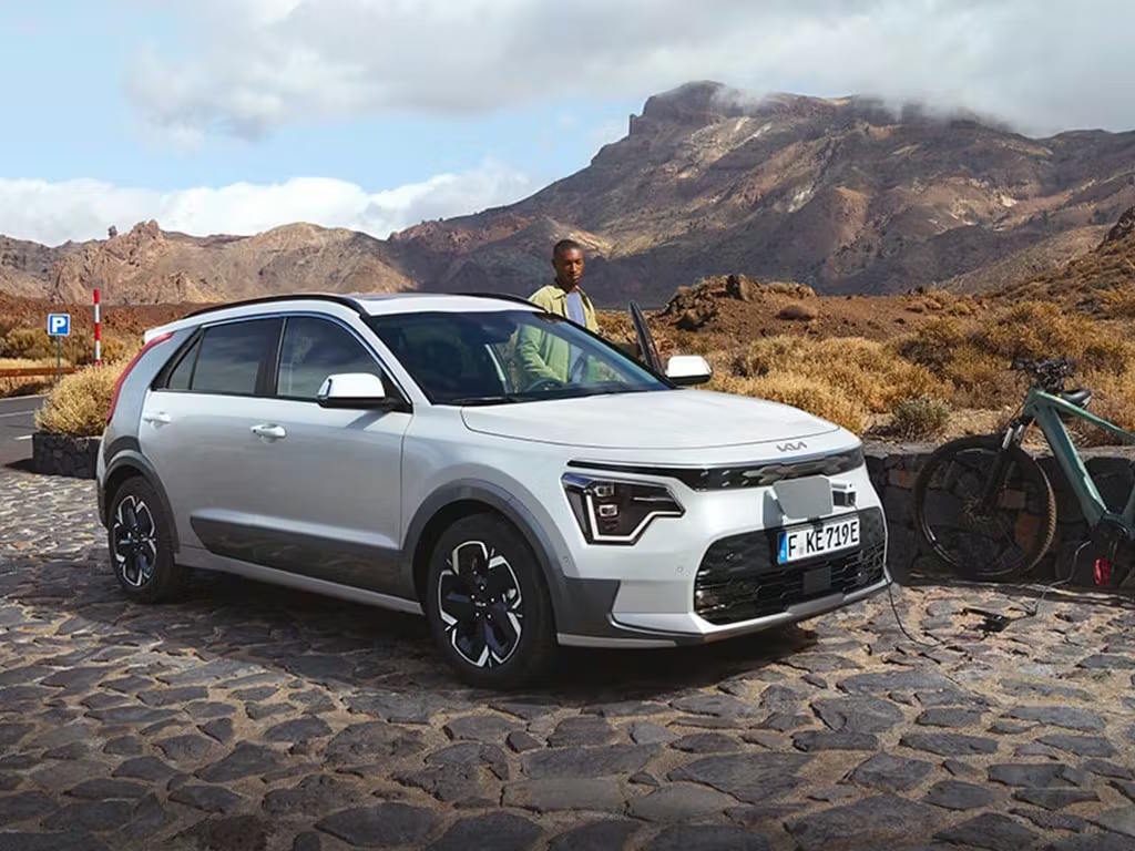 Front three-quarter view of the Kia Niro in white, featuring distinctive LED daytime running lights, black contrasting roof and alloy wheels, parked against a dramatic mountain backdrop.