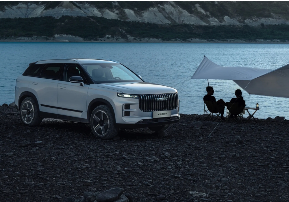 White Jaecoo 7 parked by a lakeshore beside a small canopy where two people sit, suggesting an outdoor camping setup.