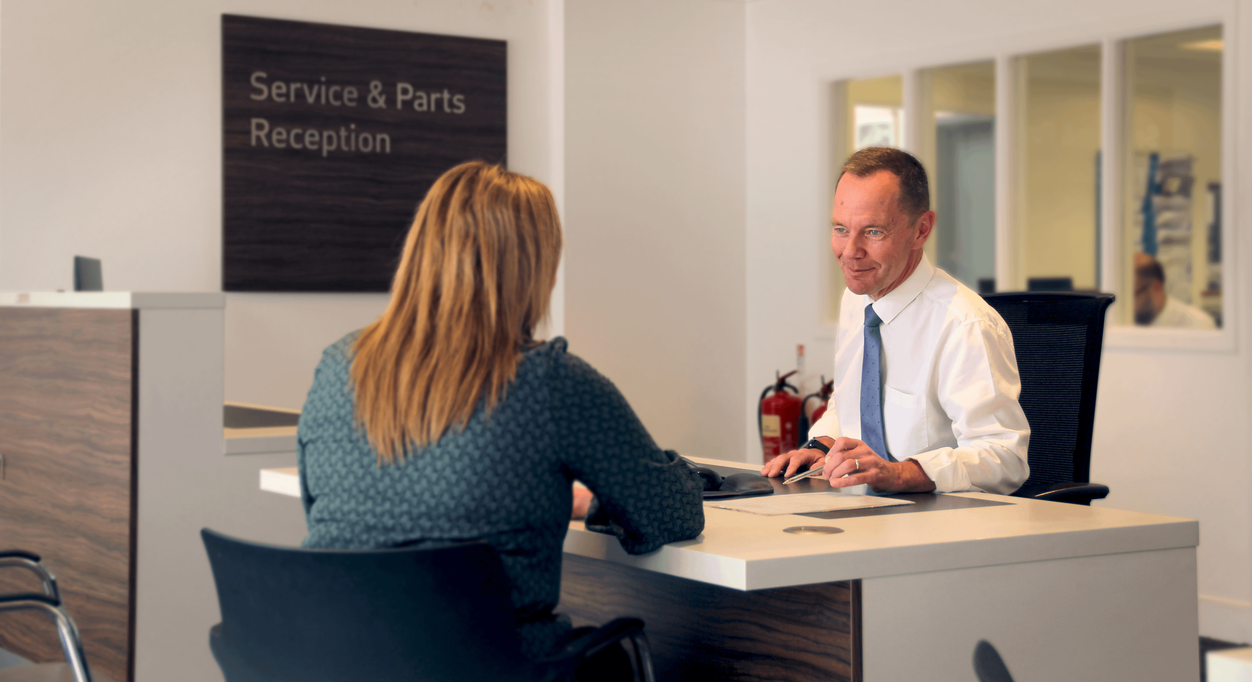 Dealership service adviser in white shirt and tie assisting a female customer at the Service & Parts Reception desk in a modern, professional showroom environment.