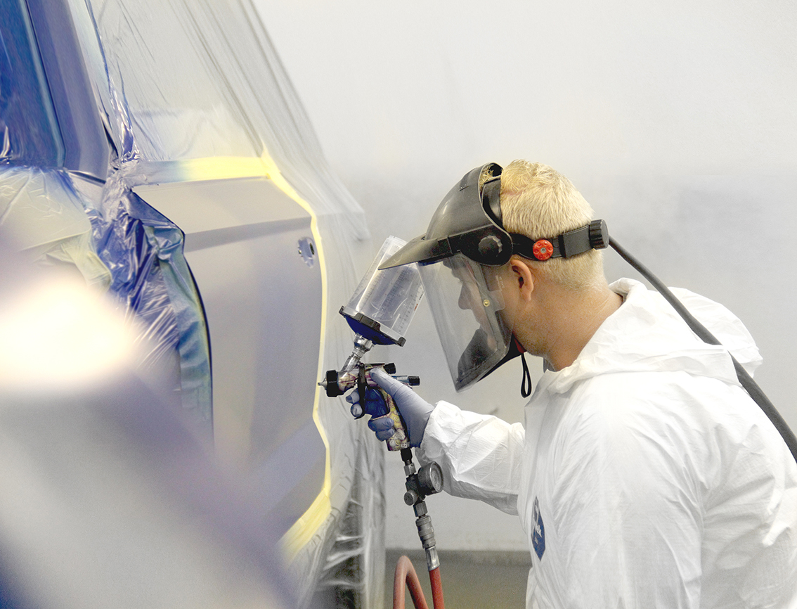 Skilled technician in protective gear applying paint to a vehicle body panel using a professional spray gun in a controlled paint booth environment.