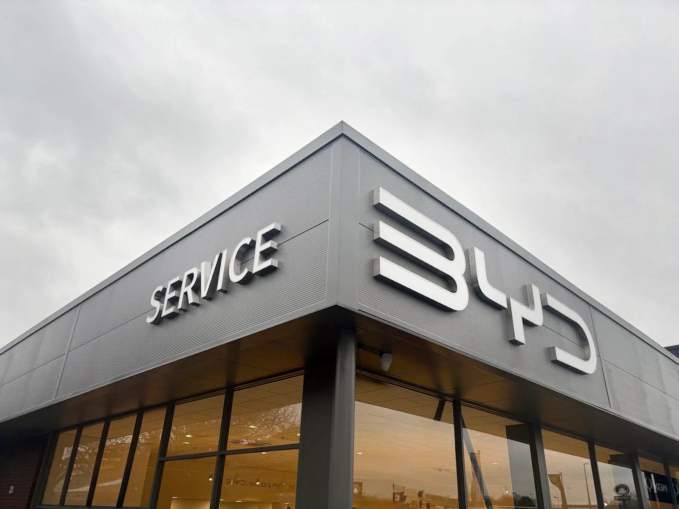 Exterior corner view of the BYD Bolton dealership, featuring modern grey cladding, illuminated BYD logo and prominent Service signage above full-height glazed showroom windows.