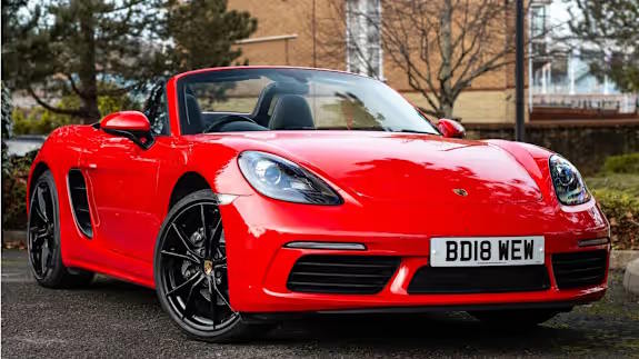 Red Porsche 718 Boxster convertible with black wheels, roof down, parked on tarmac in front of trees and buildings, viewed from the front three‑quarter angle.