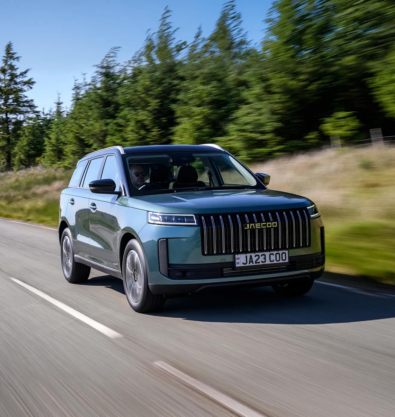 Front three-quarter view of the Jaecoo J7 SUV in motion, featuring its bold vertical grille design, slim LED headlights and sophisticated teal grey metallic finish on a country road.