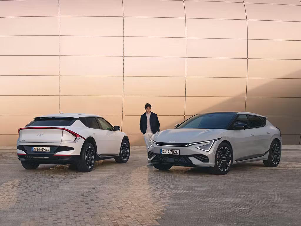 Two Kia EV6 electric crossovers parked facing opposite directions, showcasing the distinctive rear light bar design and sharp front LED daytime running lights against a copper-toned backdrop.