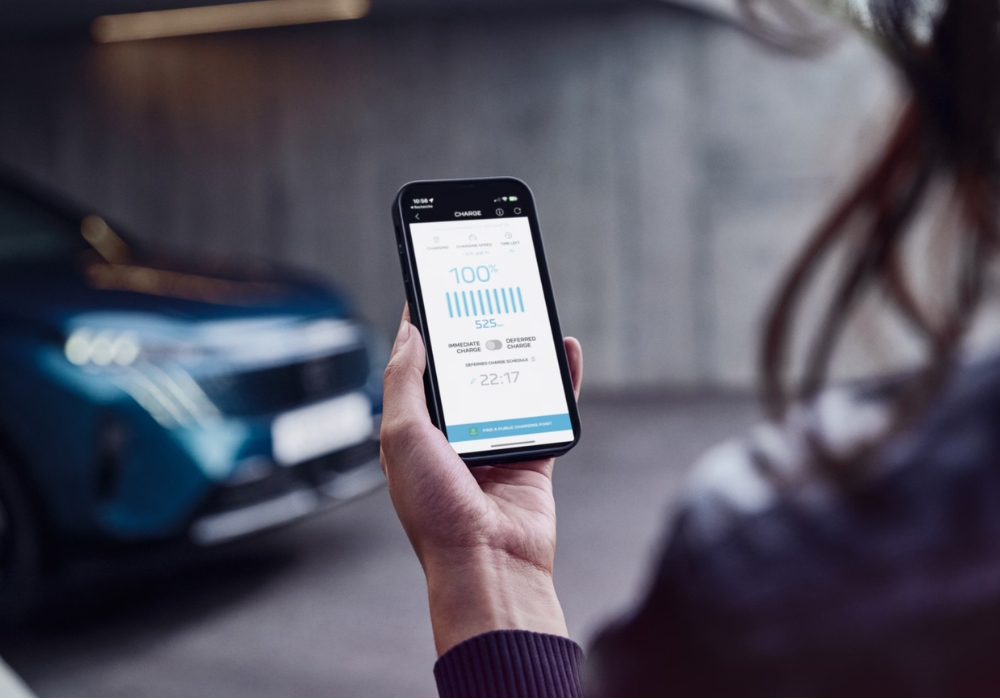 Close-up of a smartphone displaying Peugeot charging app with battery status and range information, car blurred in background.