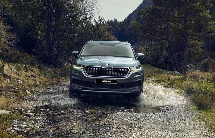 Skoda Kodiaq driving through a shallow rocky stream surrounded by trees and mountains.
