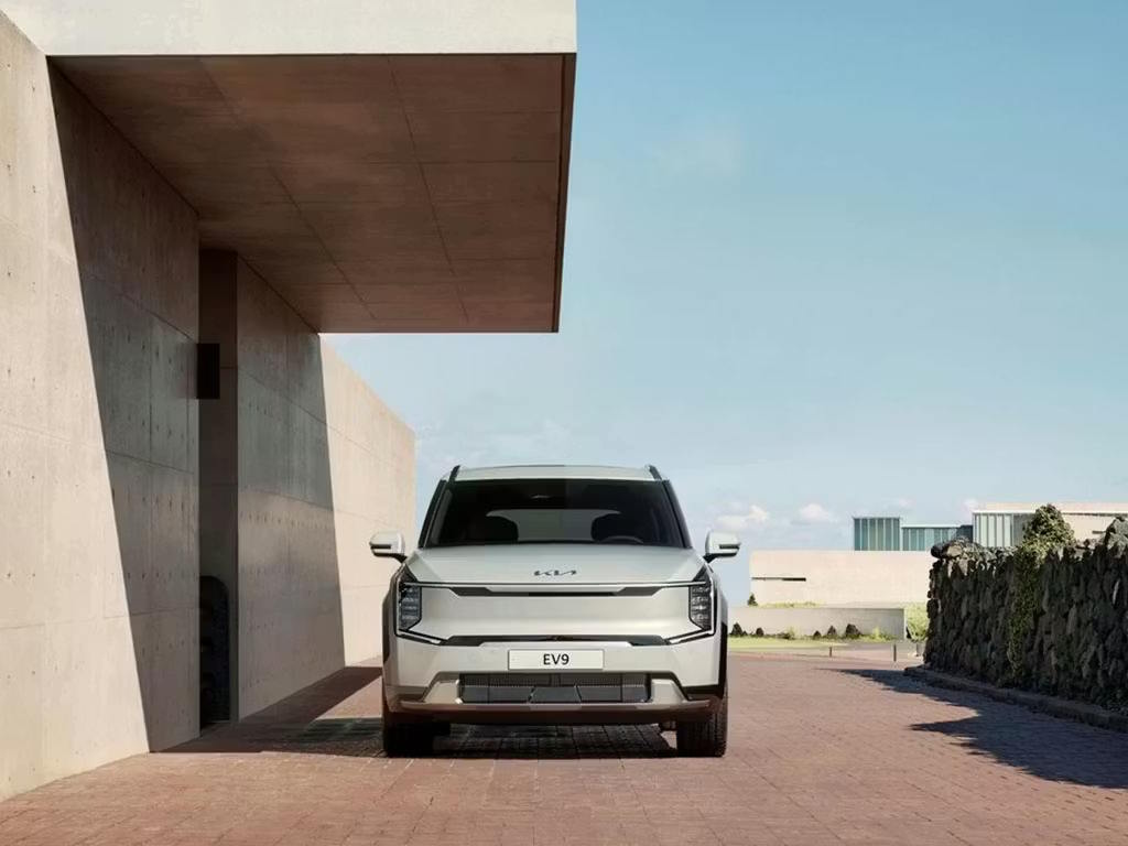 Front view of the Kia EV9 electric SUV in white, showcasing its bold geometric grille, distinctive vertical LED headlights and commanding presence against modern architecture.