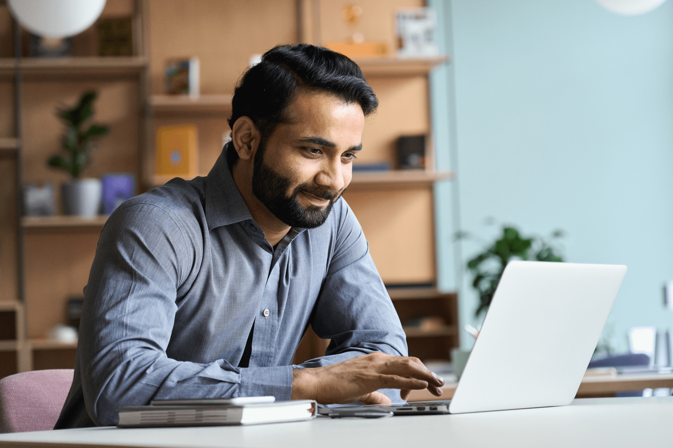 Man wearing light blue/grey shirt whilst using a laptop with a notebook and phone by his side.